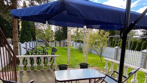 Eine Terrasse im Freien mit Tisch und Stühlen, einem blauen Sonnenschirm und einem weißen Zaun im Hintergrund.