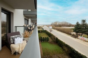 Ein Balkon mit Blick auf eine Straße und Grünflächen, eine Bank und ein kleines Gebäude in der Ferne.
