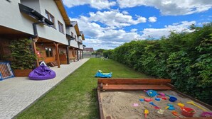 A courtyard with a wooden play area, a grassy lawn, and a building with multiple windows and a sloped roof. - Terraced Home in Sarbinowo Near the Beach (Mielno)