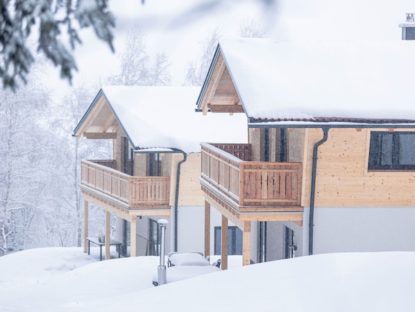 A wooden cabin with a balcony and a covered porch, surrounded by snow.