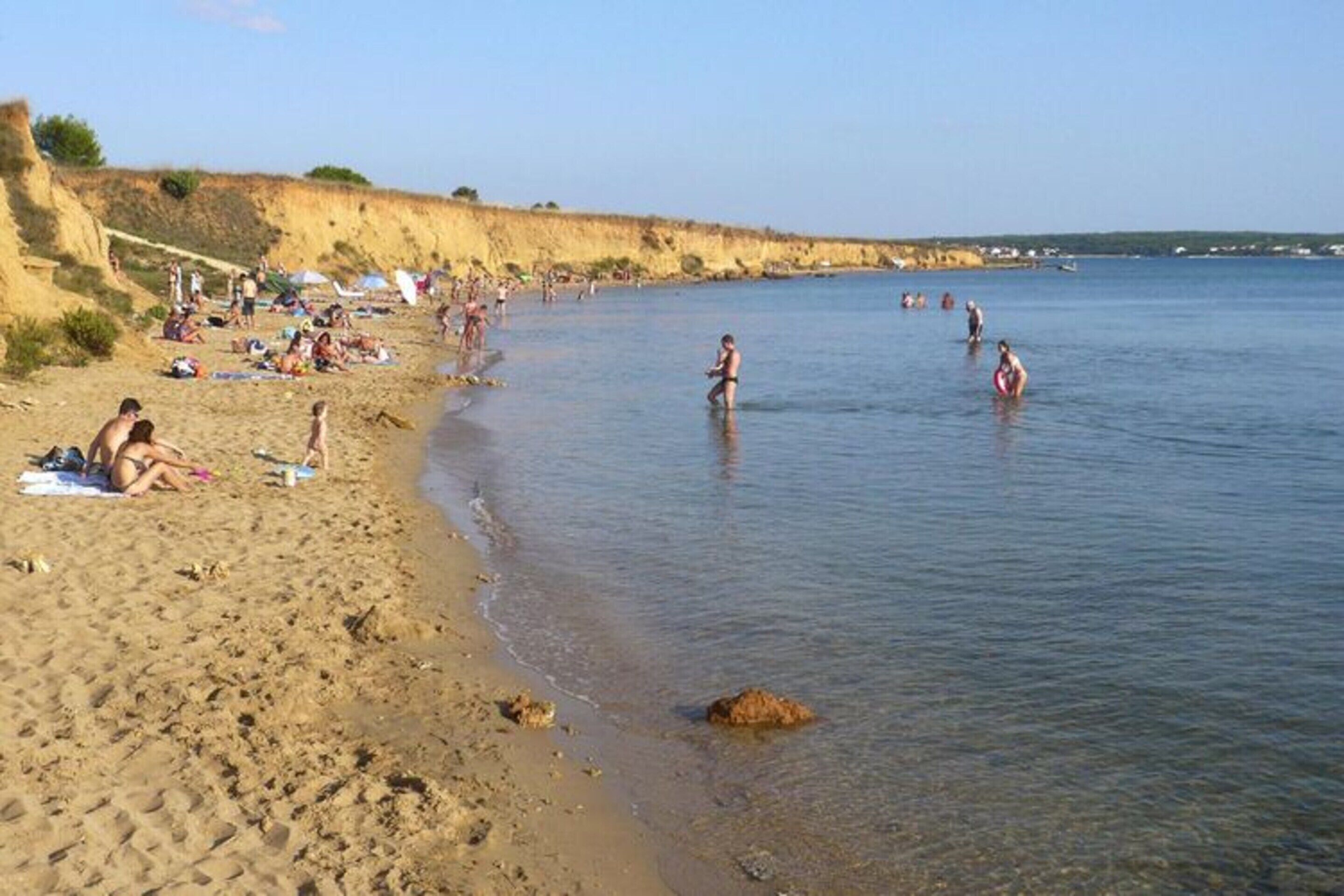 Una platja amb persones prenent el sol i banyant-se, aigua cristal·lina i sorra daurada.