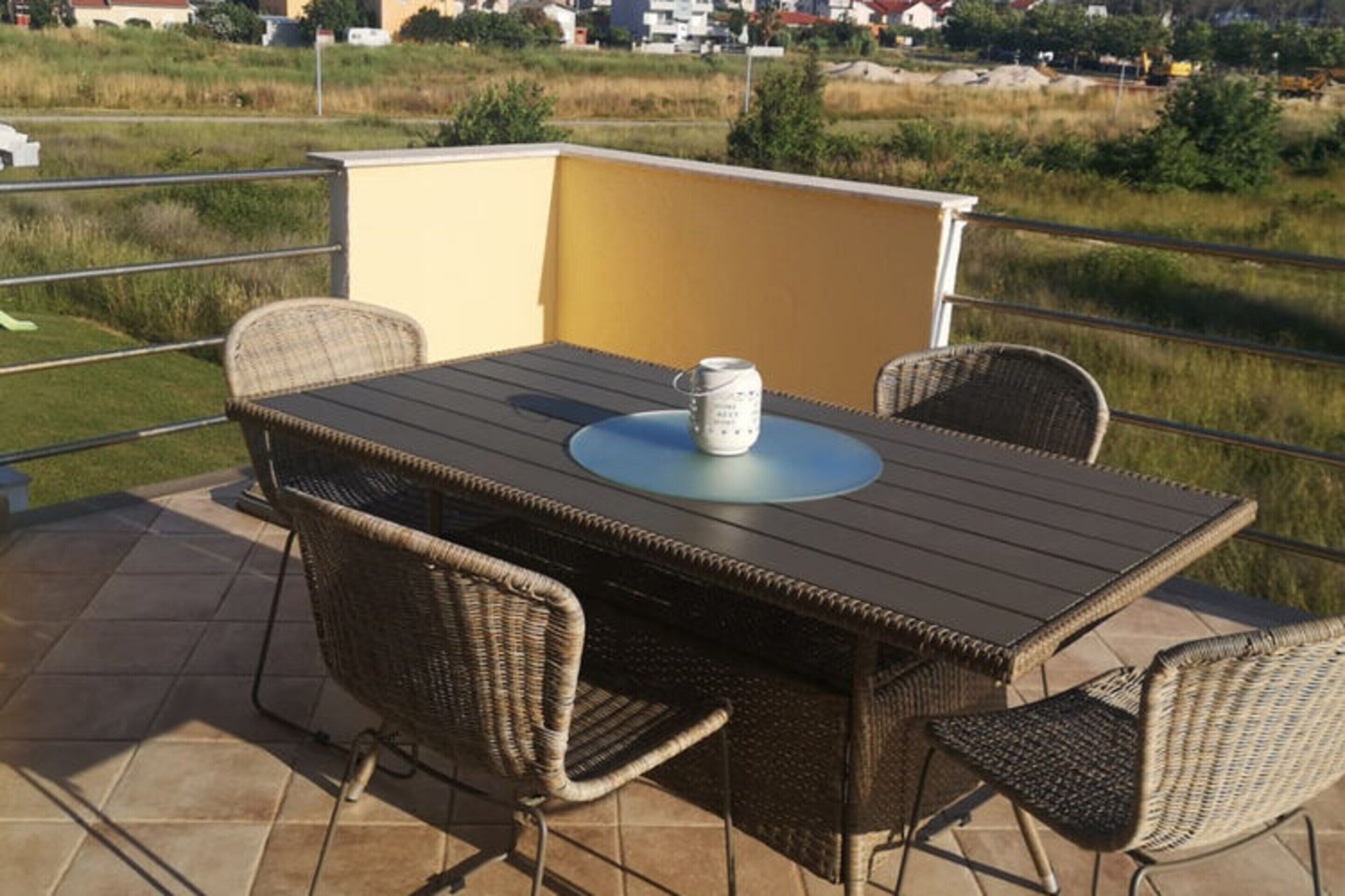 Eine Terrasse mit Tisch und Stühlen, einer kleinen Tischlampe und Blick auf ein Wiesenfeld sowie Gebäude im Hintergrund.