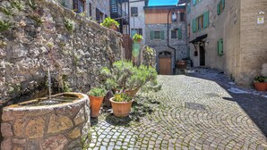 Una strada acciottolata con pareti in pietra, piante in vaso e una piccola fontana.