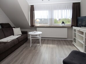 A living room with a brown sofa, a white coffee table, a TV on a stand, and a window with curtains. - Ferienwohnung am Deich in Berumbur (Berumbur)
