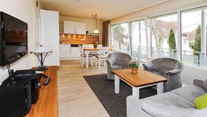 A modern living room with a flat-screen TV, a wooden coffee table, and grey armchairs. A kitchen with white cabinets and a dining area with a table and chairs is visible in the background. - Residenz Seestern, Boltenhagen (Boltenhagen)