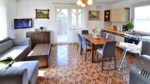 Een moderne woonkamer met een bank, salontafel en een eethoek met stoelen. Er is een televisie aan de muur bevestigd, een keuken met een fornuis en kastjes, en een groot raam.