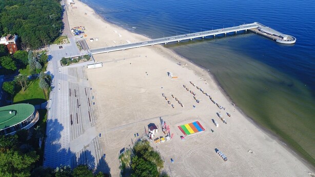 Luchtbeeld van een strand met een lange pier die het water in reikt, een zandstrand met een vlag in opvallende kleuren, en een gebouw met een groen dak.