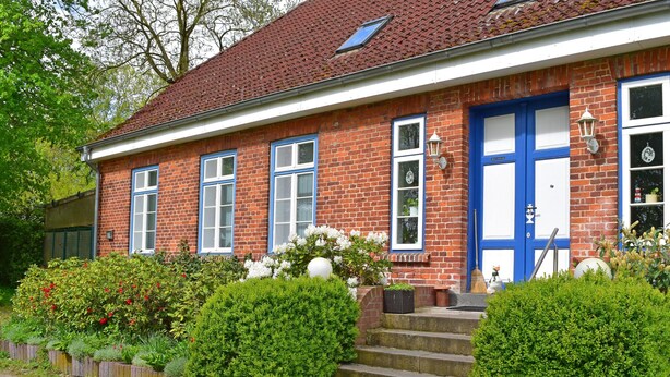 Une maison traditionnelle en briques, avec un toit recouvert de tuiles rouges, des fenêtres à cadre blanc et une porte bleue.