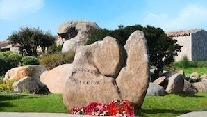 A large rock with "RESIDENCE Punta Falcone" inscribed on it, surrounded by a garden with flowers and greenery.