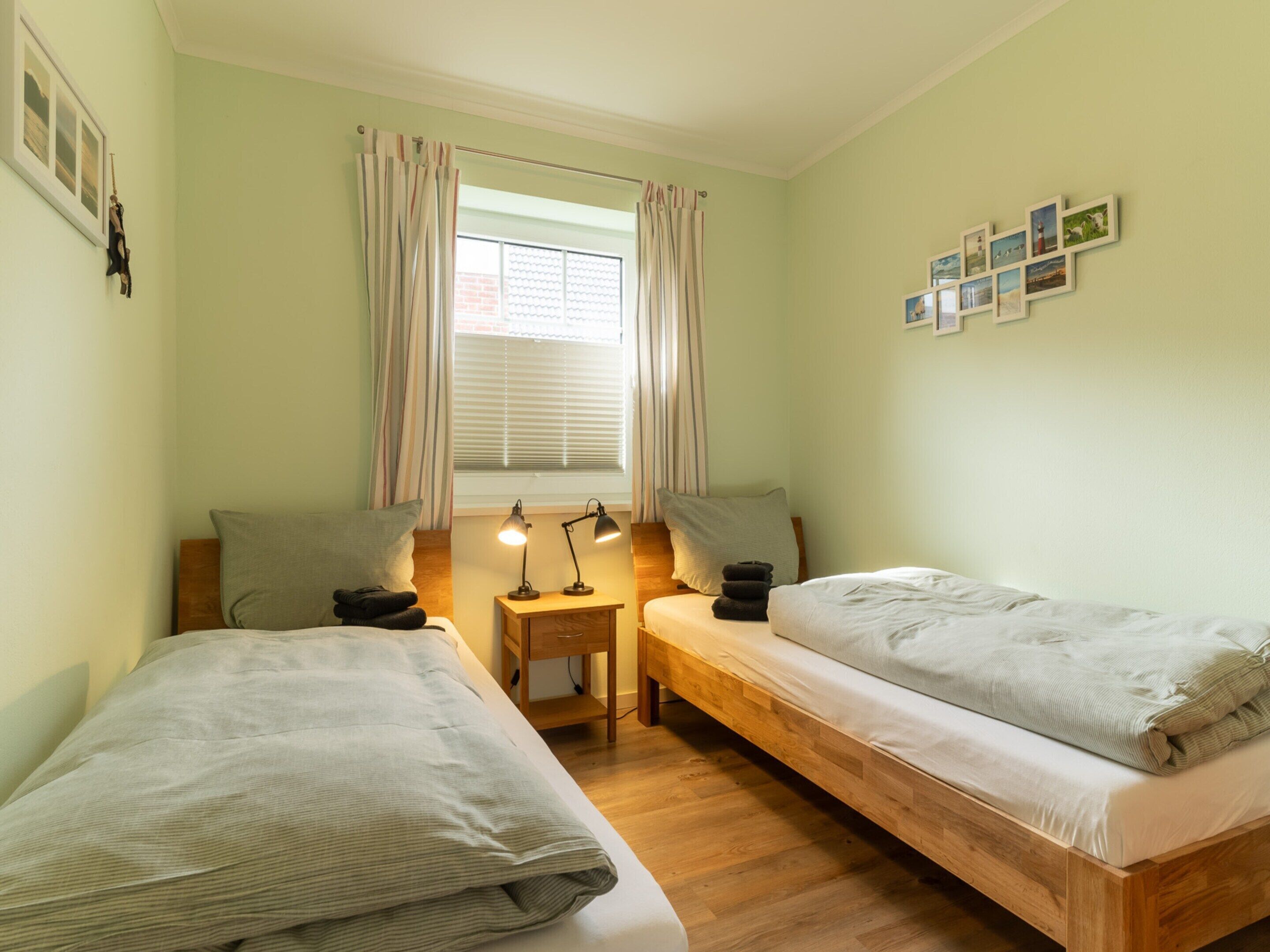 Une chambre avec deux lits simples, une petite table de chevet avec des lampes, une fenêtre avec des rideaux et un mur décoré de tableaux encadrés.