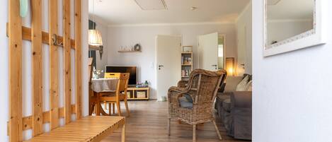 Un salón amplio con un banco de madera, un sillón de mimbre y un sofá. Un televisor está sobre un mueble bajo, y hay una estantería con libros y objetos decorativos.