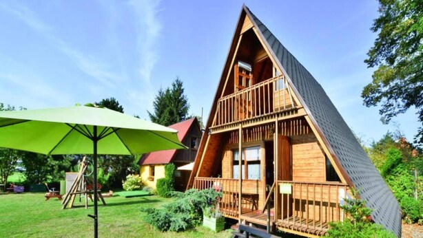 A wooden A-frame cabin with a deck, surrounded by a well-maintained lawn and trees. - Comfortable Holiday Home Near Lake Jamno (gmina Sianów)