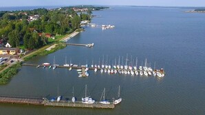 An aerial view of a marina with numerous boats docked, a pier extending into the water, and a town with houses and greenery in the background. - Comfortable Holiday Home Near Lake Jamno (gmina Sianów)