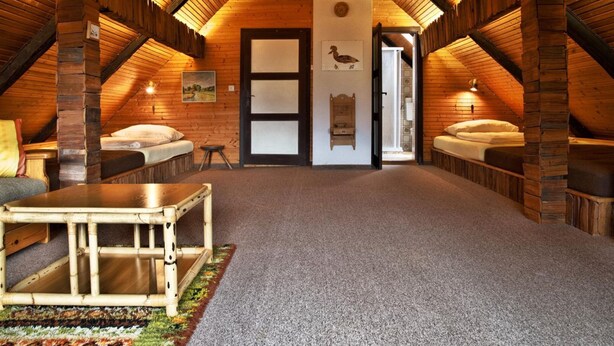 Une chambre mansardée chaleureuse avec un plafond en pente, des poutres en bois, deux lits, une petite table et une porte donnant sur une autre pièce.