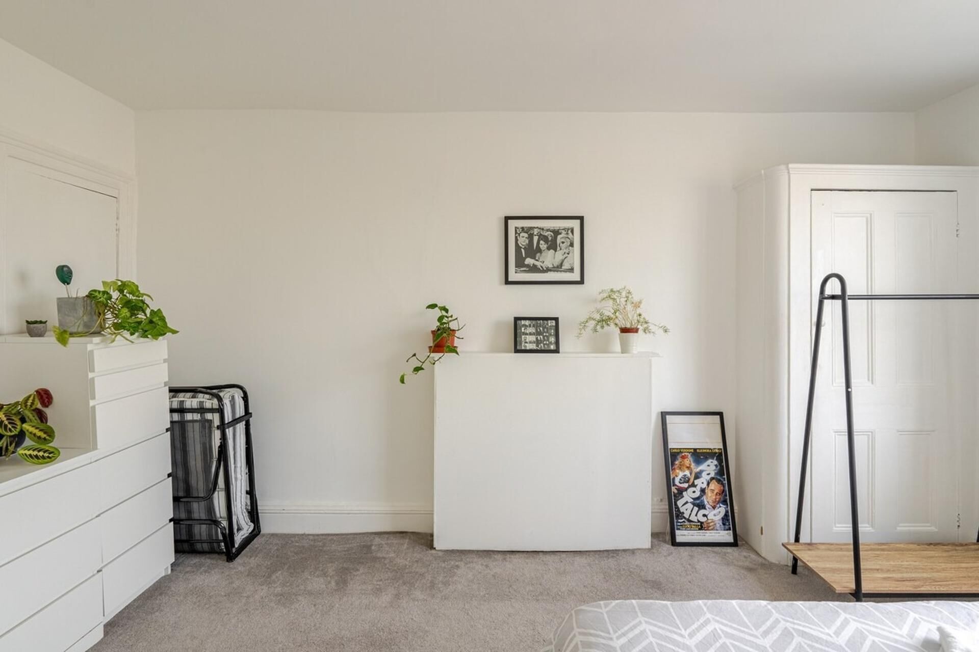 A minimalist bedroom with a white wardrobe, a small desk, and a bed with a patterned bedspread.