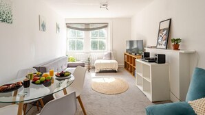 A modern living room with a glass dining table, a sofa, a TV, and a bookshelf. - London Oasis Retreat - 2 Bedroom Apartment (London)
