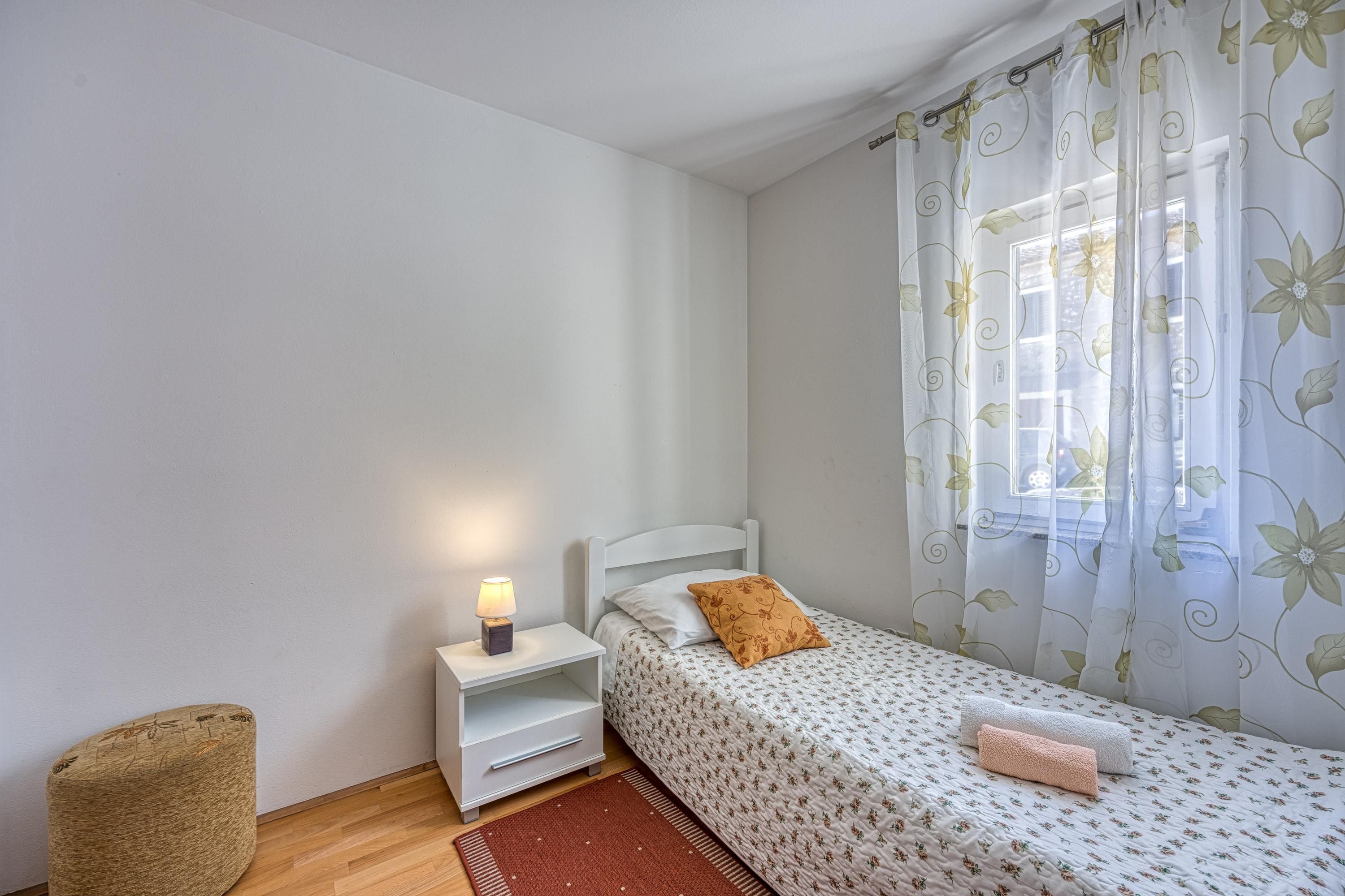 A small, neatly arranged bedroom with a single bed, a nightstand with a lamp, a small chair, and a window with floral curtains.