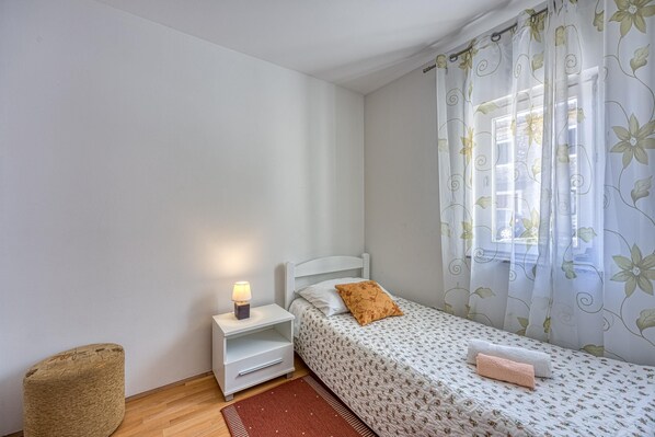 Une petite chambre à coucher, bien rangée, avec un lit simple, une table de chevet avec une lampe, une petite chaise et une fenêtre ornée de rideaux à motifs floraux.