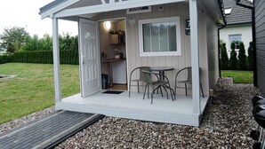 A small, white, single-story structure with a covered patio area, a door, a window, and a table with chairs. - Dziwnowek Holiday House Near Beach (Kamien Pomorski)