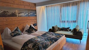 Et soveværelse med sengegavl i træ, seng med blomstret sengetøj, sofa med puder og et vindue med lette gardiner.