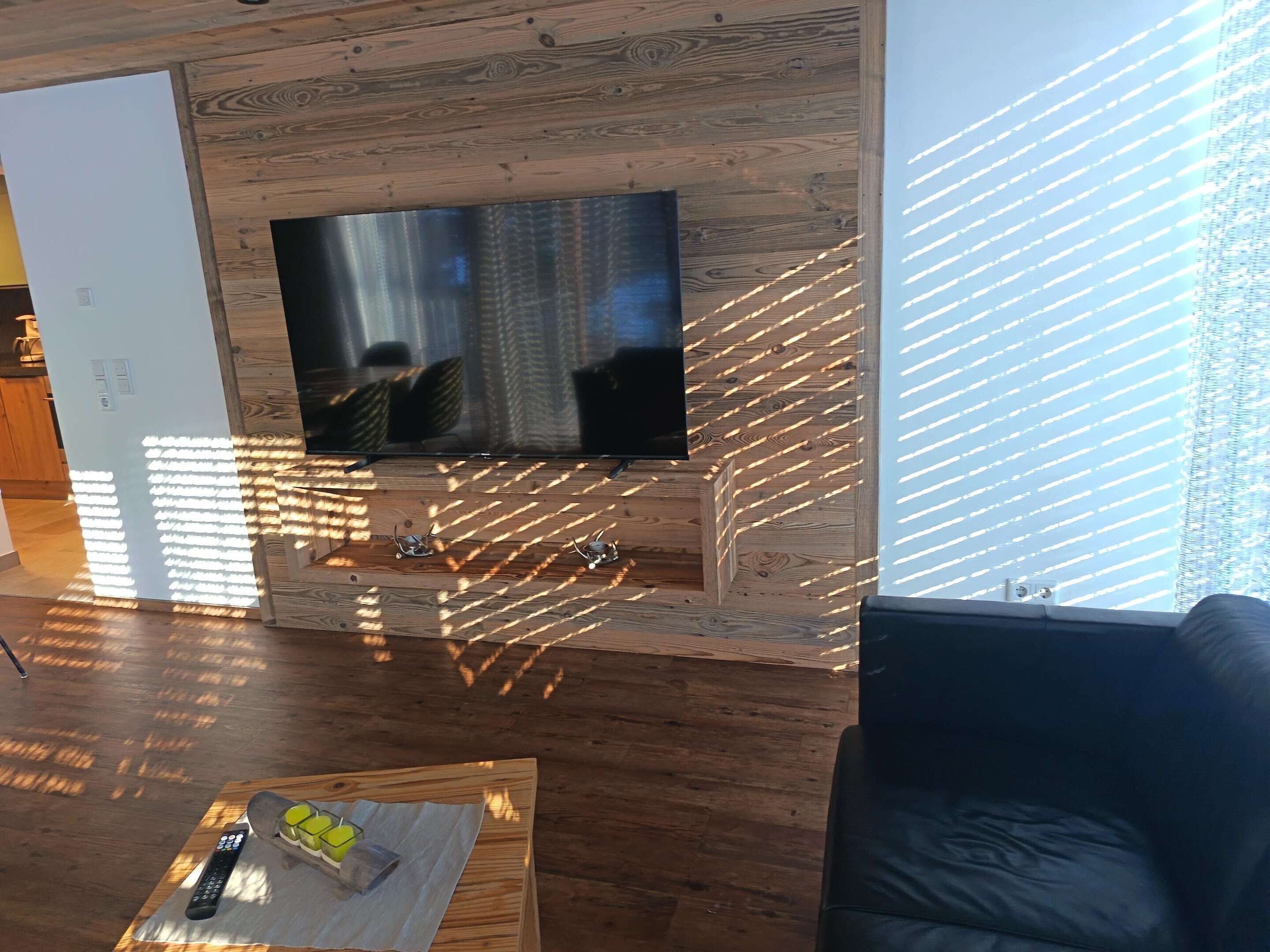A living room with a wooden wall, a flat-screen TV mounted on it, a coffee table with a remote control and a glass, and a black leather couch.