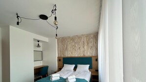 A modern hotel room with a teal bed, a wooden headboard, a desk with a chair, and a hanging light fixture. - Apartment for 5 People Near the sea in Grzybowo (Kolobrzeg)