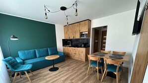 A modern living space with a teal sofa, a round wooden table, and a kitchen area with wooden cabinets and a microwave. - Apartment for 5 People Near the sea in Grzybowo (Kolobrzeg)