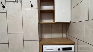 A compact laundry room with a front-loading washing machine, a wooden shelf above, and a tiled wall. - Apartment for 5 People Near the sea in Grzybowo (Kolobrzeg)