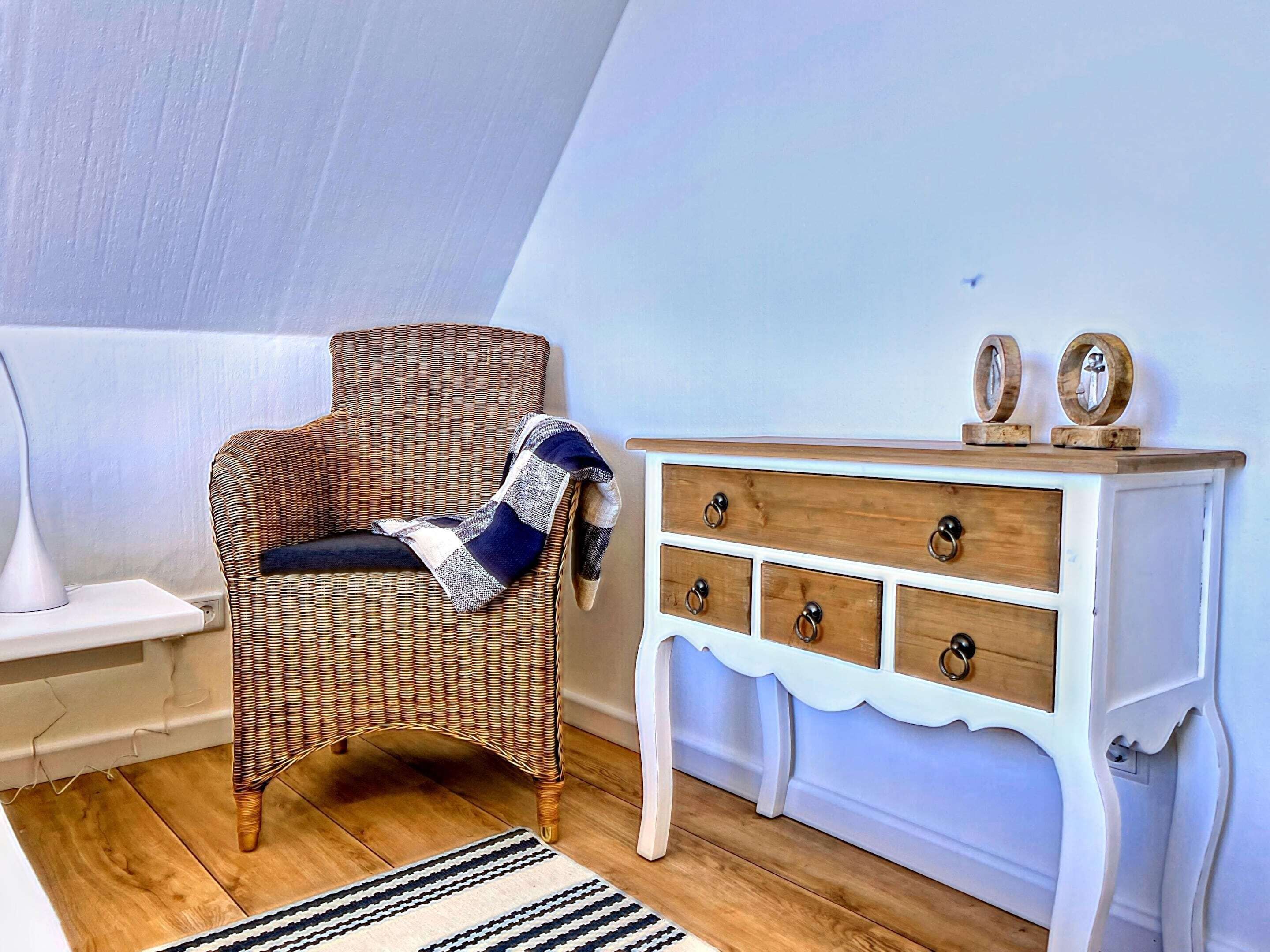 A corner room with a wicker chair, a white dresser with wooden knobs, and a striped rug on a wooden floor.