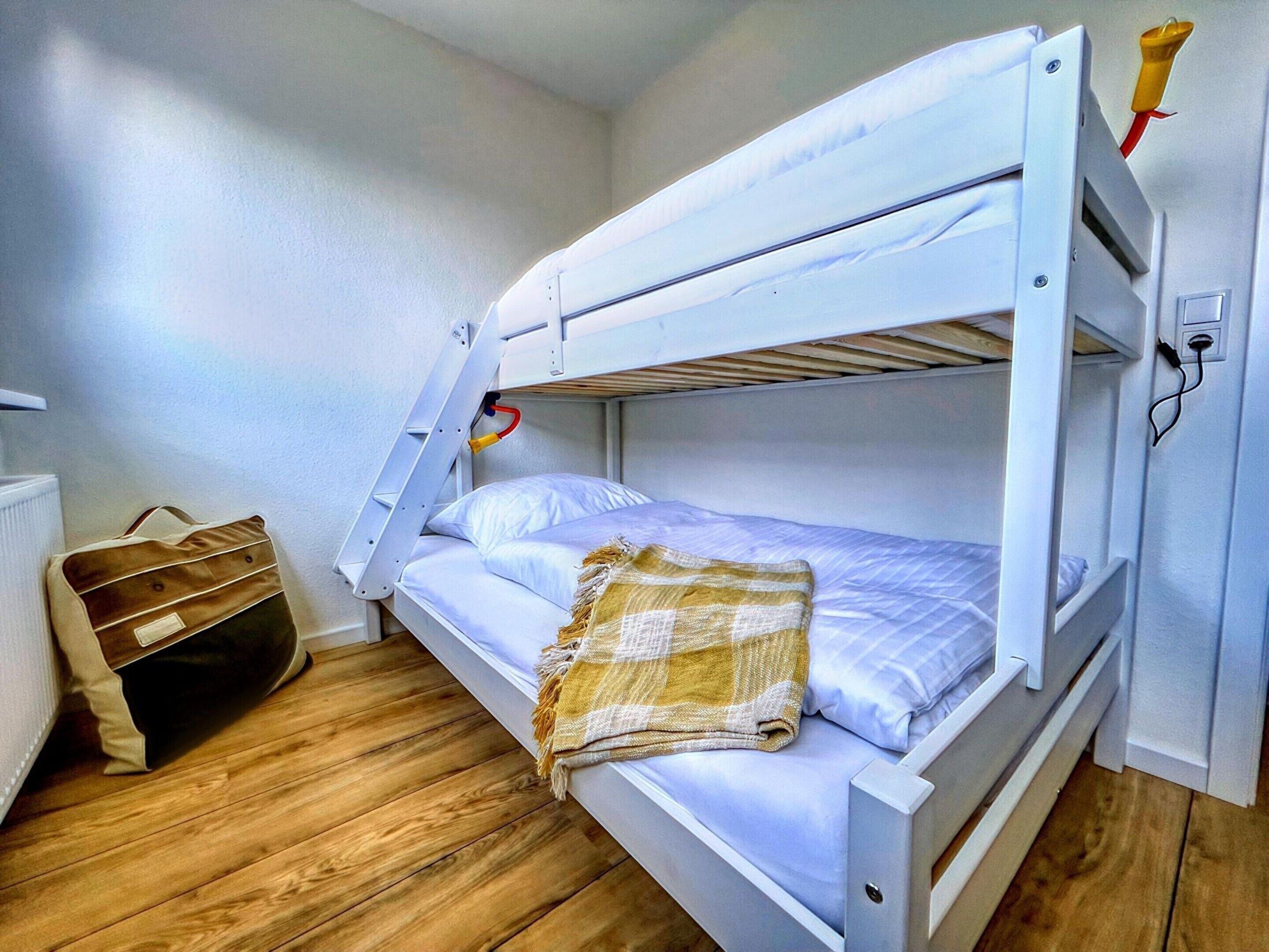 Un lit superposé avec une échelle, un plancher en bois, un radiateur et un sac posé sur le sol.