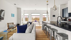 A modern kitchen with a black countertop, stainless steel appliances, and a bar area. There is a dining area with a wooden table and chairs, and a living room with a sofa and a coffee table. - Gorgeous New Build ~Luxury Finishes~ Huge Balcony (Austin)