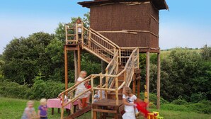 A wooden play structure with stairs and a platform, surrounded by greenery.