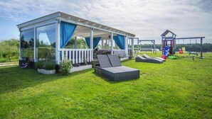 A covered outdoor seating area with a glass enclosure, a cushioned bench, and a playground with slides and swings. - Rewal Cottage Near the Baltic Beach (Rewal)