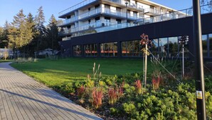 Un immeuble moderne à plusieurs étages, doté de balcons, entouré d’un jardin bien entretenu et bordé d’une allée pavée.