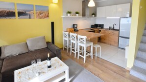 A compact living space with a kitchenette, a dining area with white chairs, a sofa, and a coffee table with a bottle and glasses. - Cozy Cottage for 2 People Near the Sandy Beach (gmina Bedzino)