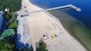 An aerial view of a beach with a long pier extending into the water, a sandy beach area with umbrellas, and a building with a green roof. - Holiday Home in Rewal Near Baltic Beach (Rewal)