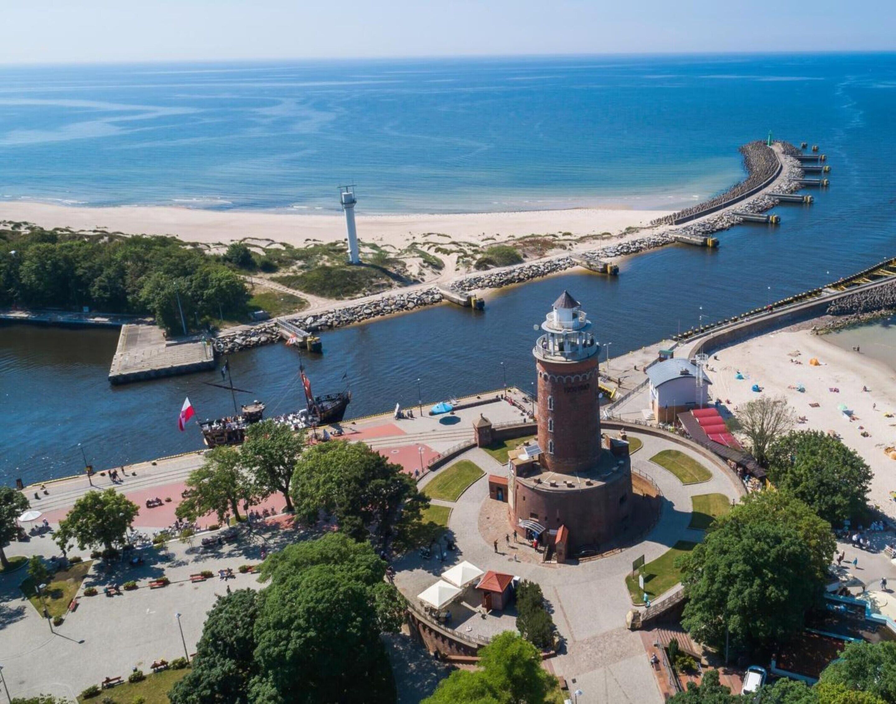 A coastal area with a lighthouse, a pier extending into the water, and a beach with people and umbrellas.