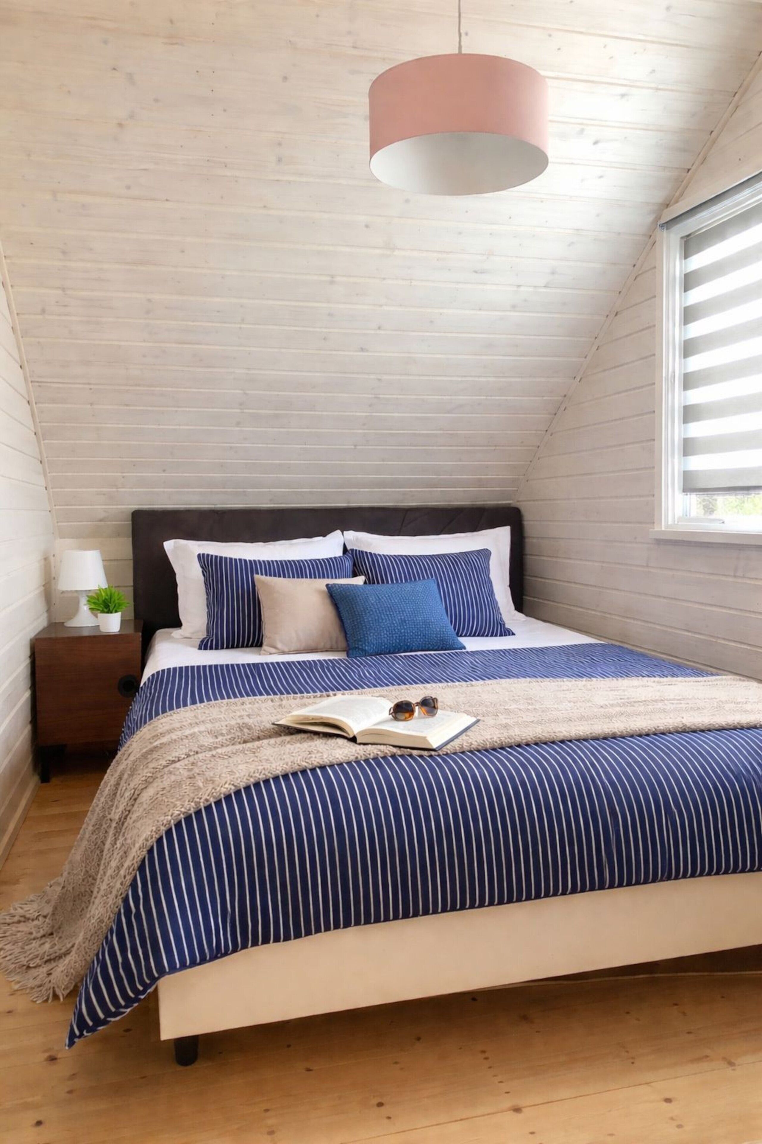 Ein Schlafzimmer mit schräg angelegter Decke, einem Bett mit gestreifter Bettwäsche, einem Nachttisch mit Lampe und einem Fenster mit Jalousien.