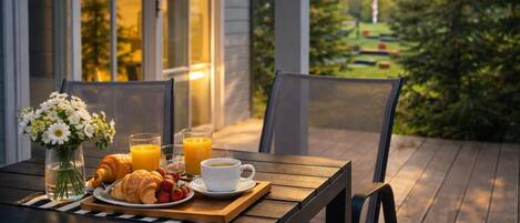 Un desayuno dispuesto en una terraza de madera con una mesa, sillas, un jarrón de flores, croissants, fresas, una taza de café y vasos de zumo de naranja.