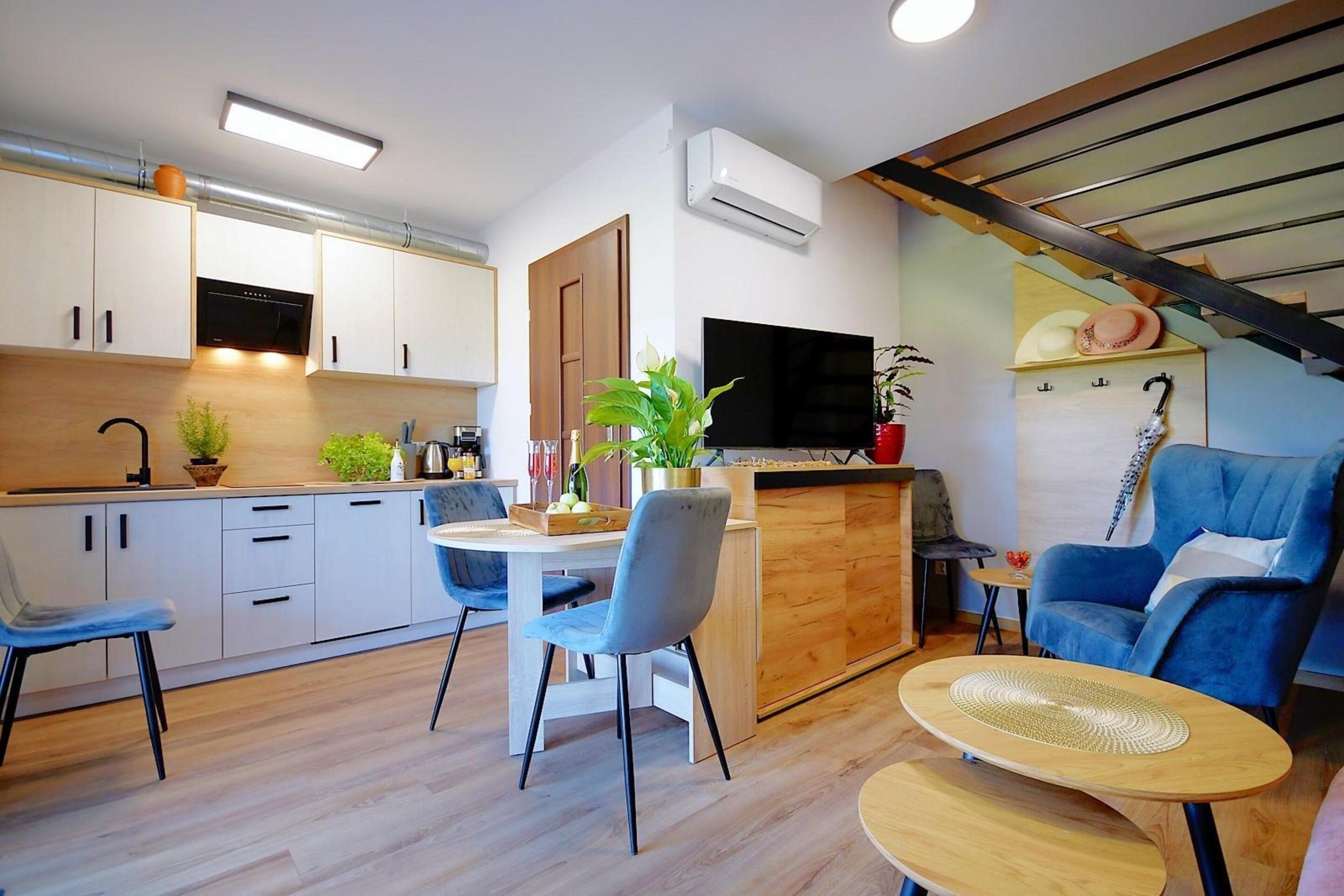 Una cucina moderna con mobili bianchi, una zona pranzo con sedie blu, un televisore montato a parete e una scala con ringhiera in legno.