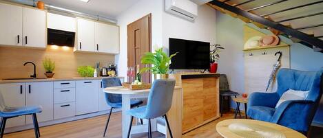 A modern kitchen with white cabinets, a dining area with blue chairs, a TV mounted on the wall, and a staircase with a wooden railing.