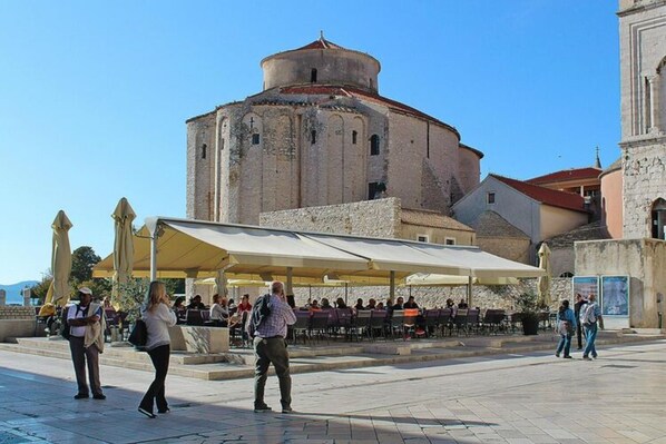 An outdoor seating area with a covered patio, a historic building with a dome in the background, and a clear blue sky.