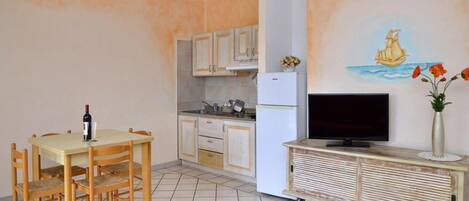Cocina compacta con mesa, sillas, refrigerador y televisor sobre un mueble de madera.