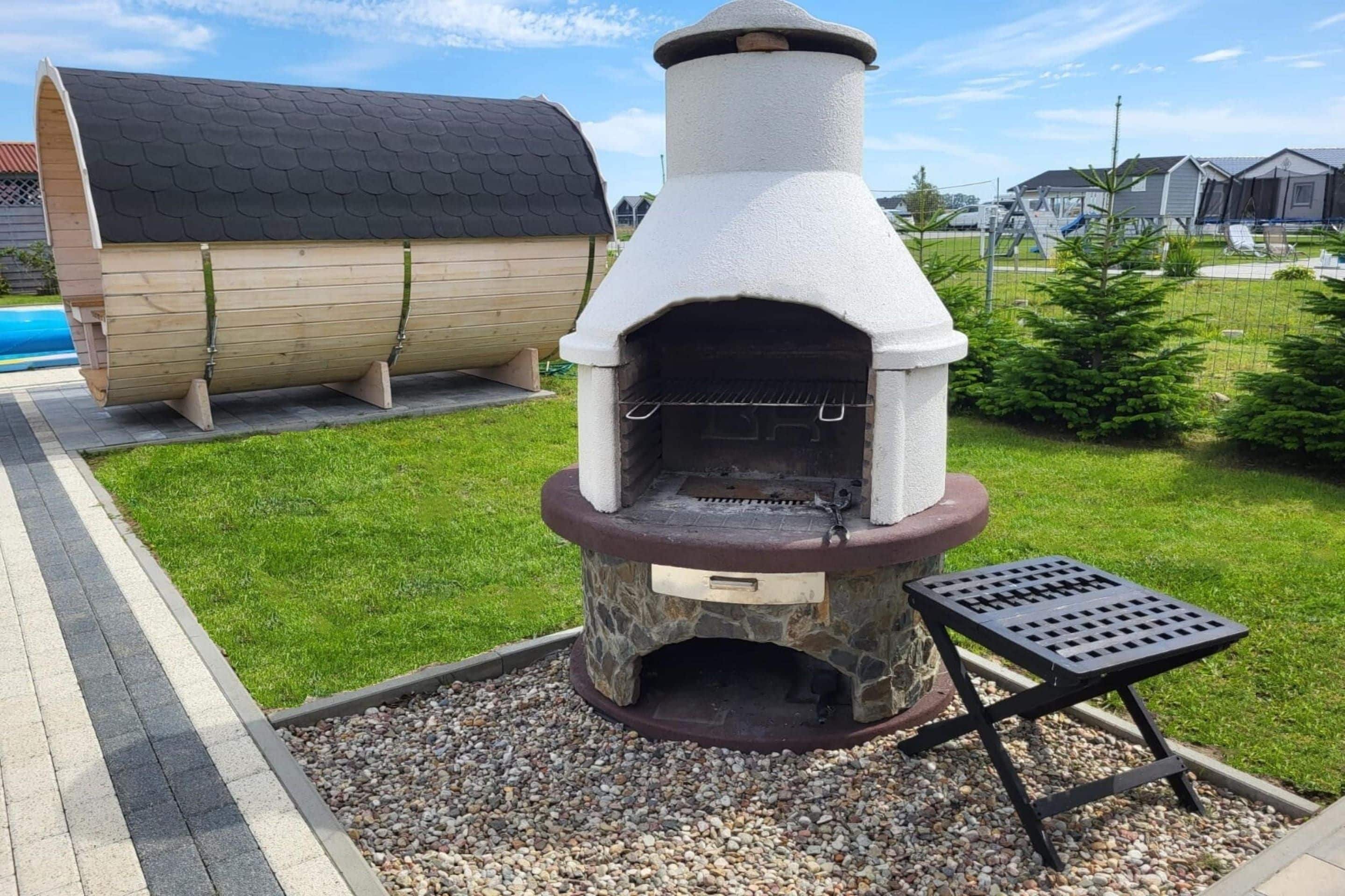 A stone-built outdoor oven with a chimney, a wooden barrel, and a metal table in a landscaped area.