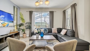 A modern living room with a sofa, coffee table, and chairs. There is a TV on the left and a large window with curtains. - EV_4958 - 4BR / 3BA Storey Lake Townhome near to Disney, Private Pool & Resort (Kissimmee)