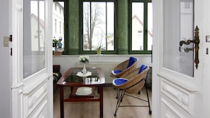 Une pièce avec une table en bois, des chaises en osier, un vase rempli de fleurs et une vue dégagée sur l’extérieur grâce à de grandes fenêtres aux montures vertes.