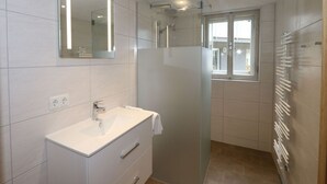 Une salle de bain moderne dotée d’une douche à parois de verre, d’un meuble-lavabo blanc avec un miroir et d’une fenêtre laissant entrer la lumière naturelle.