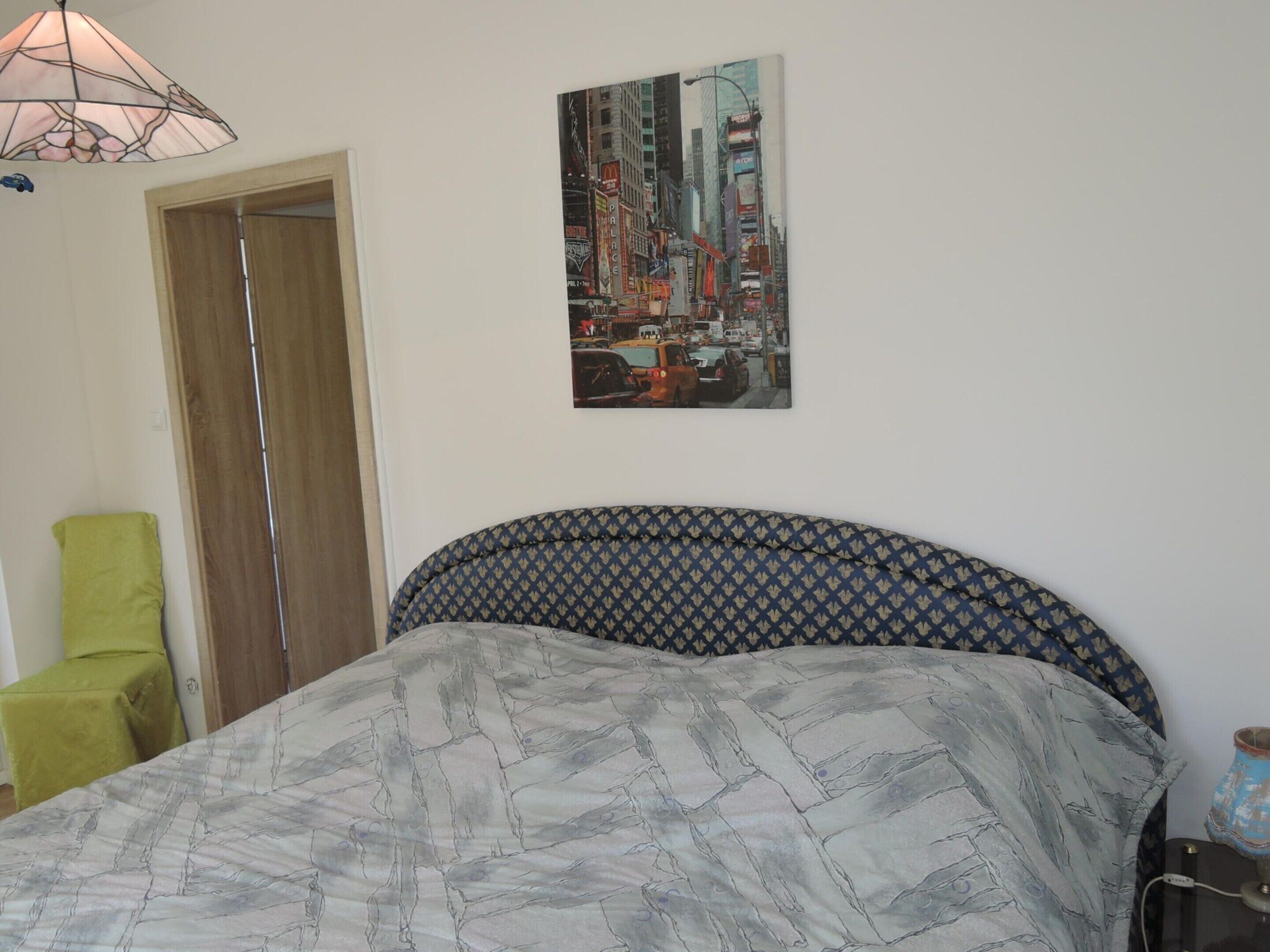Una camera da letto con un letto, una poltrona verde, un quadro che raffigura un paesaggio urbano e una lampada.