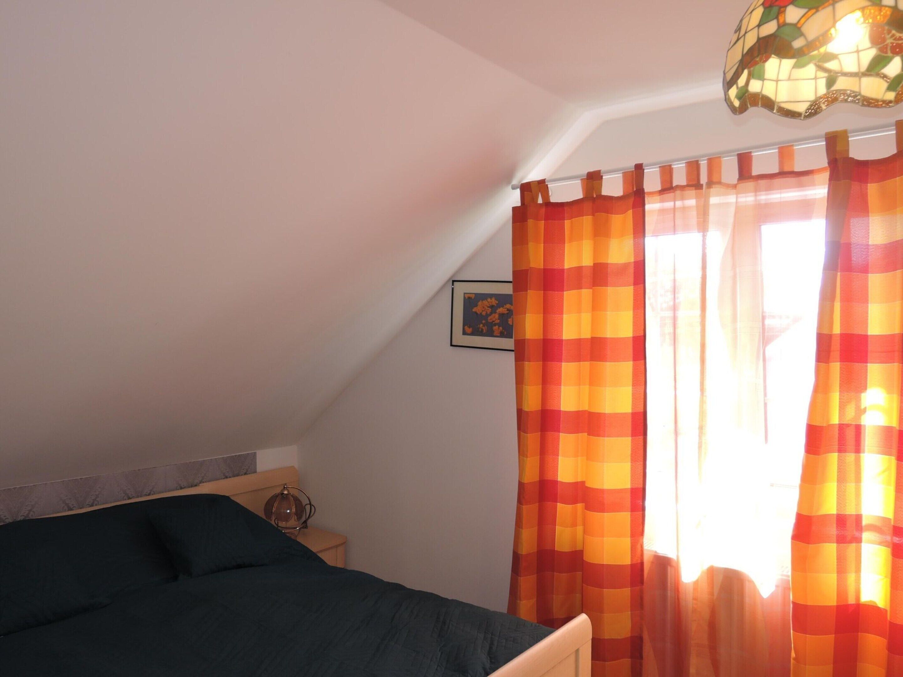 A bedroom with a bed, a nightstand, a lamp, a window with curtains, and a ceiling light fixture.