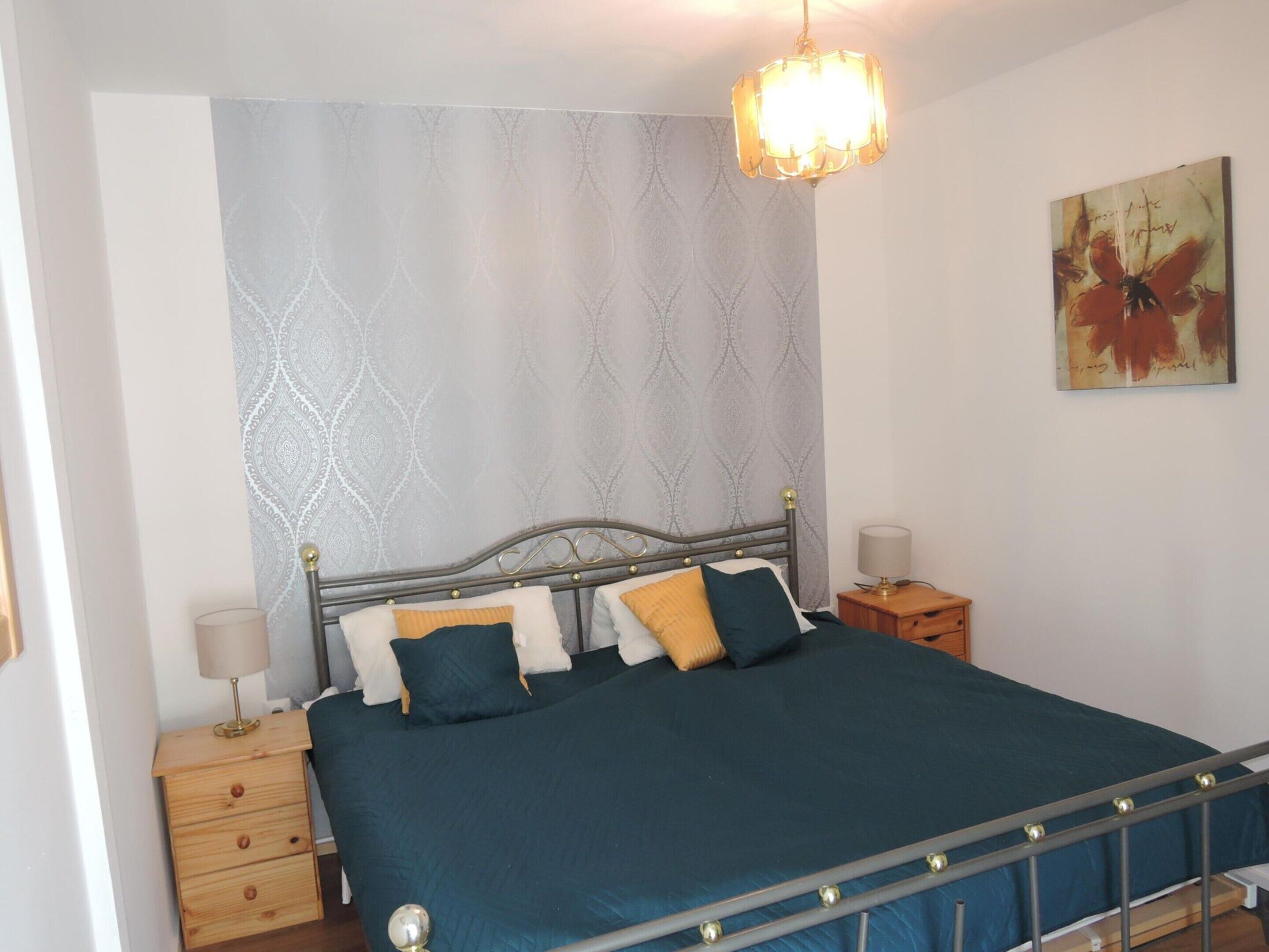 A bedroom with a metal-framed bed, a patterned wall, a painting, and a bedside lamp.